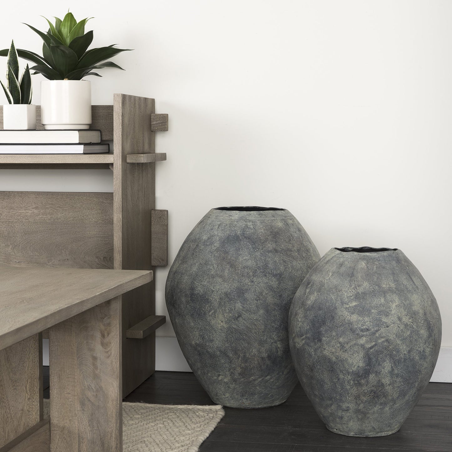 Two large gray textured vases on a dark surface with a wooden shelf and plants in the background.