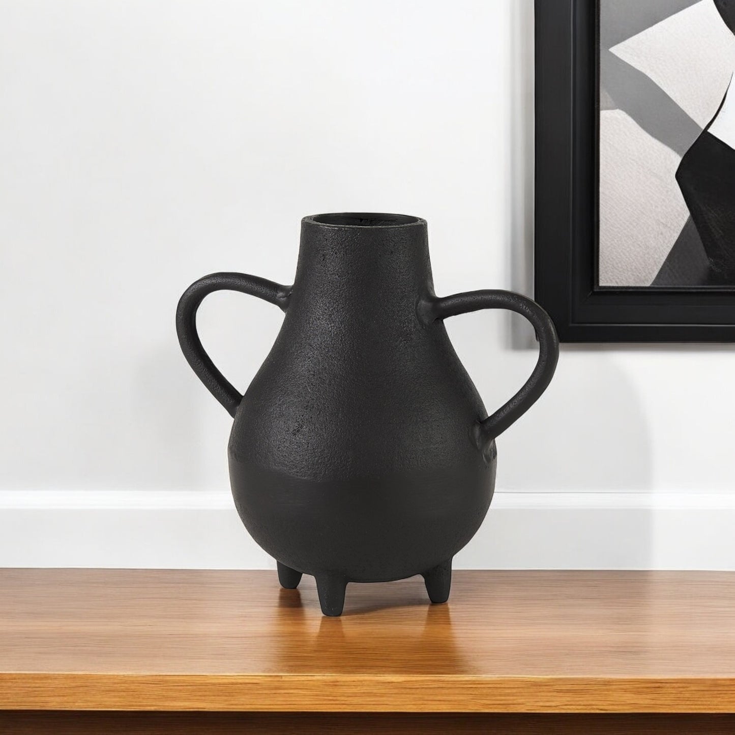 Black vase with handles on a wooden surface against a white wall with a framed picture.