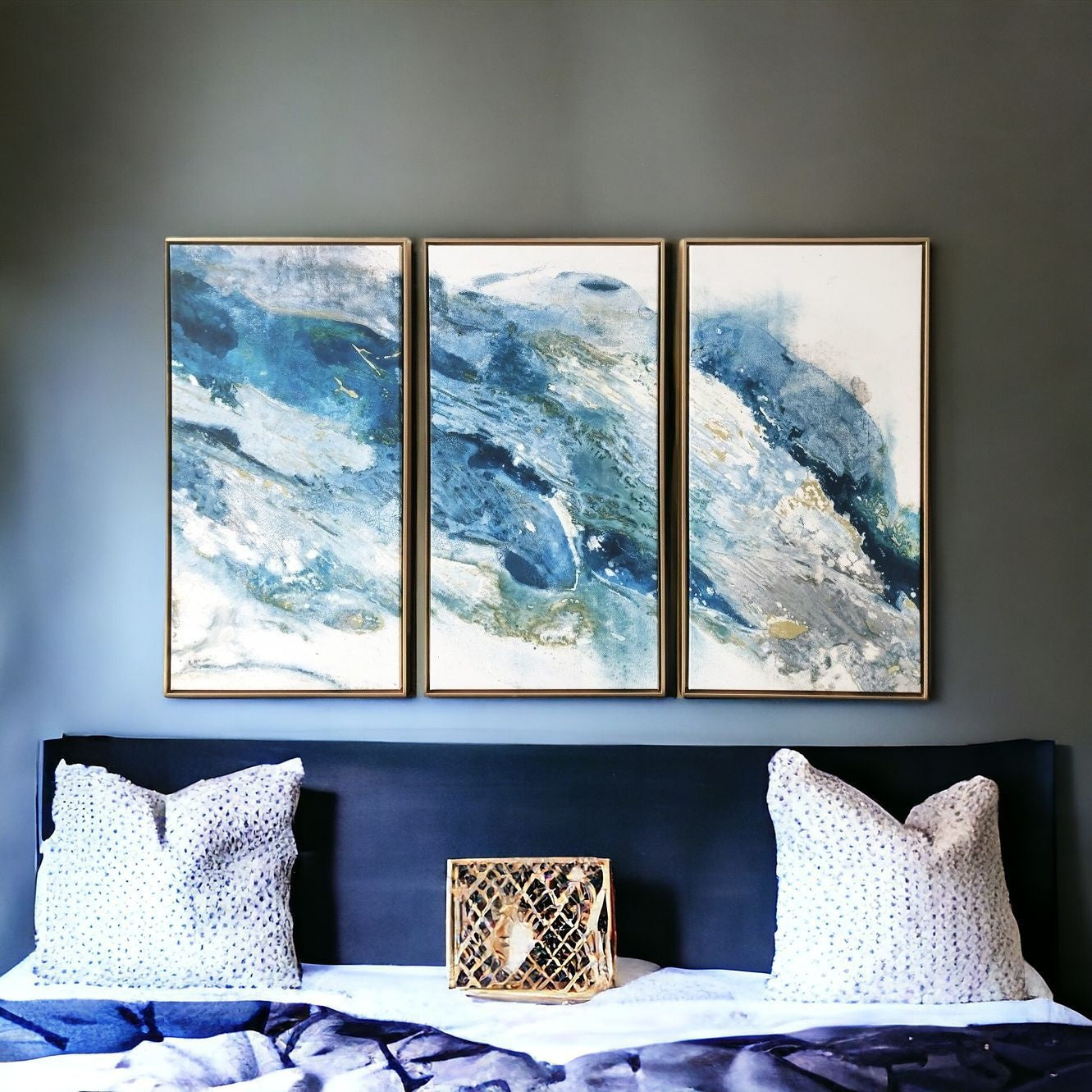 A set of three canvas wall artworks featuring abstract blue and gold colors, displayed above a bed with white bedding and two decorative pillows.