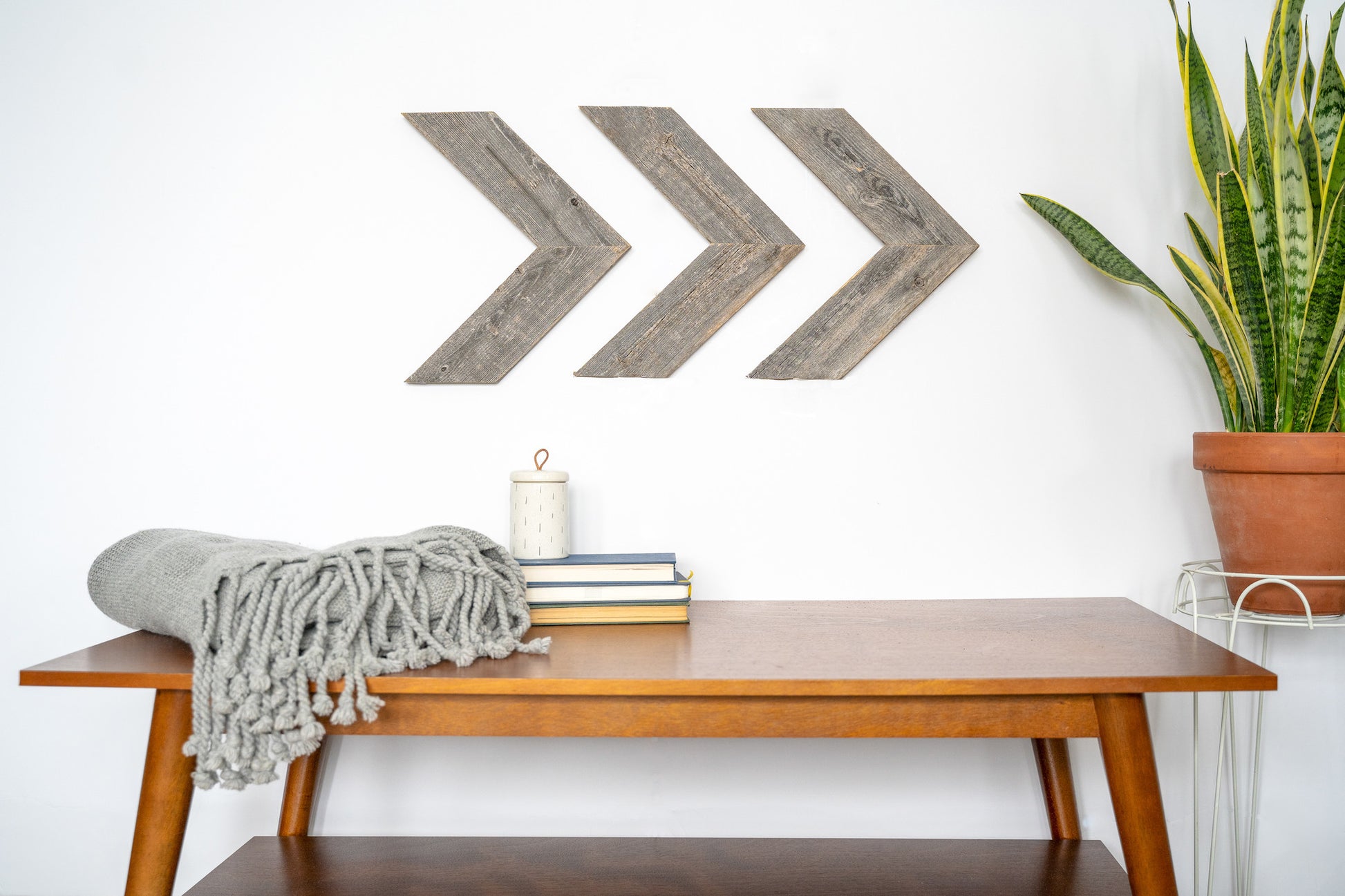 Wooden bench with a blanket, books, and a candle in front of three wooden arrow wall decorations on a white wall.