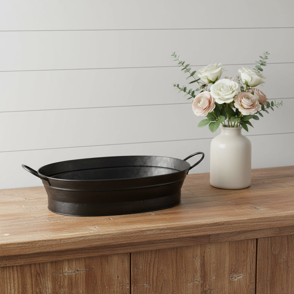 Black metal tray and white vase with flowers on a wooden surface against a white wall.