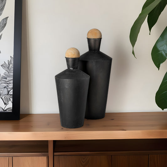 Two black ceramic bottles with wooden caps on a wooden surface against a neutral wall.