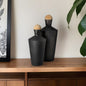 Two black ceramic bottles with wooden caps on a wooden surface against a neutral wall.