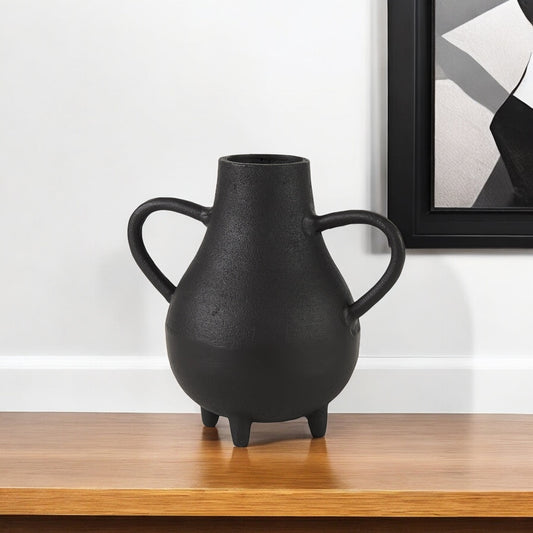 Black vase with handles on a wooden surface against a white wall with a framed picture.