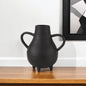Black vase with handles on a wooden surface against a white wall with a framed picture.